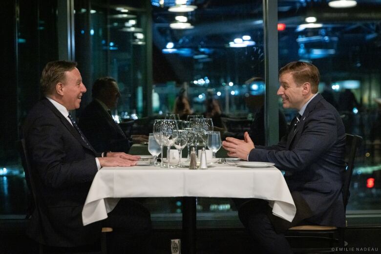 Two men in suits sit across a dinner table from each other, both smiling, with plates and wine glasses in between them.