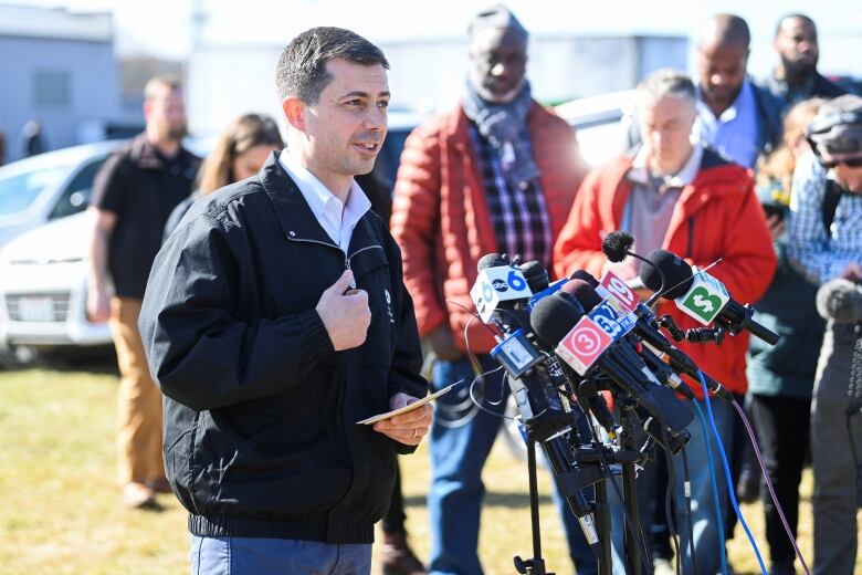 A man in a light jacket is shown speaking into microphones as others look on in the background.