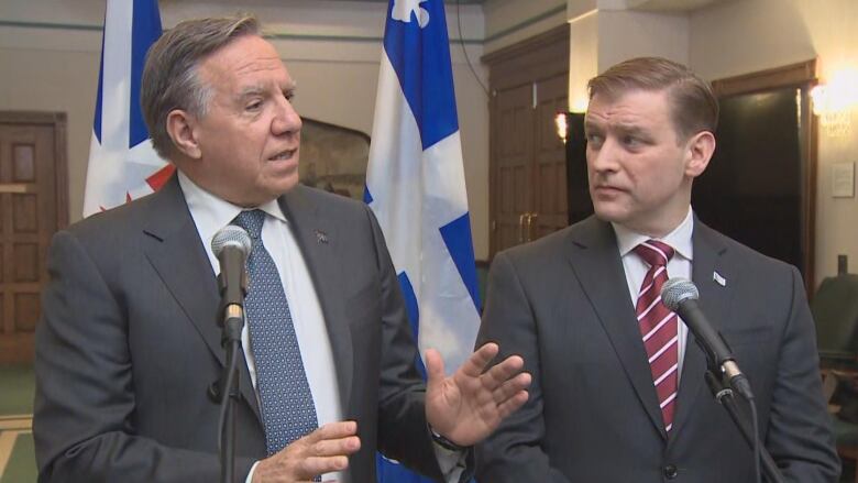 Two men in suits stand in front of microphones, while one gestures with his hands.