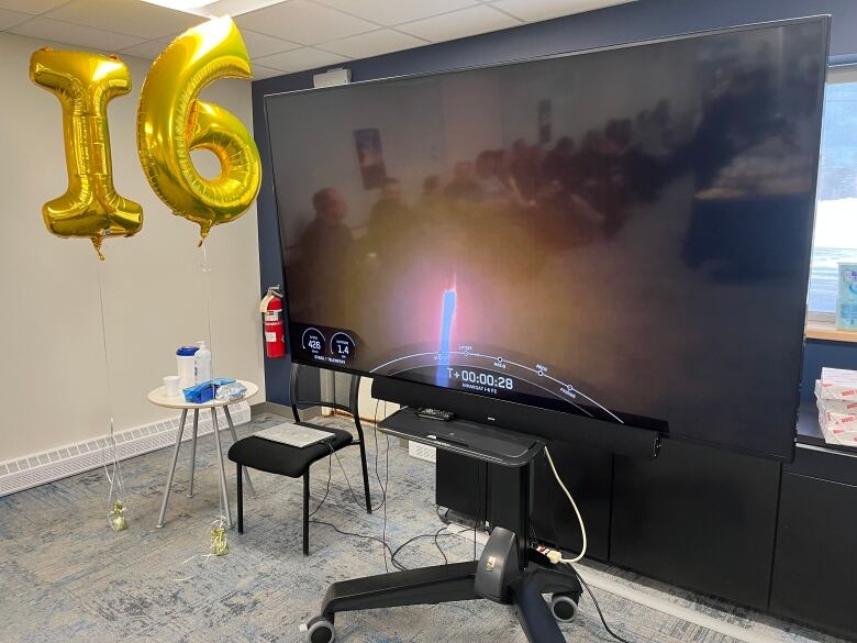 A large screen displays a rocket launch. Next to the screen are balloons that spell out I6.