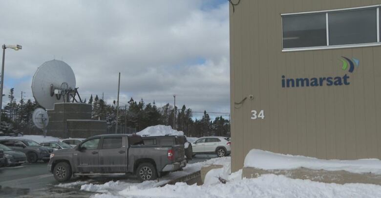 A large satellite dish sits by a snowy parking lot, outside a beige building that has the name 