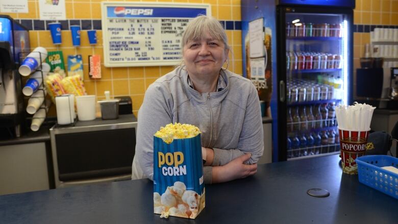 a woman in a grey sweater sits behind a movie snack counter with a bag of popcorn 