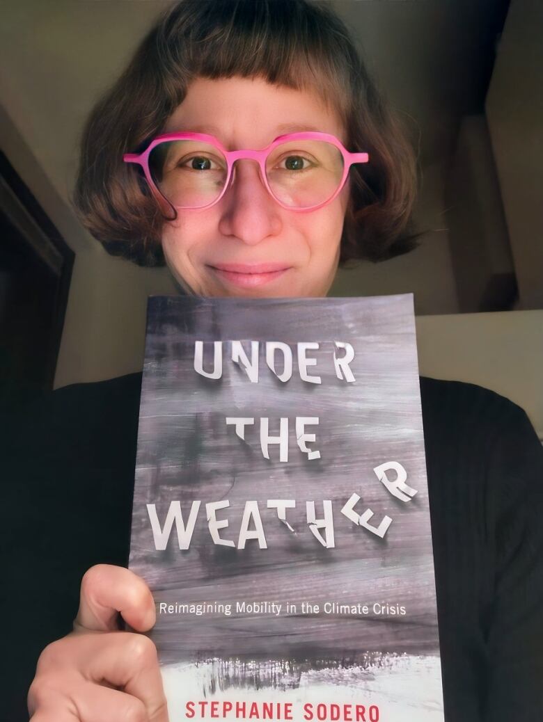 A woman wearing pink framed glasses holds up a book with the title Under the Weather.