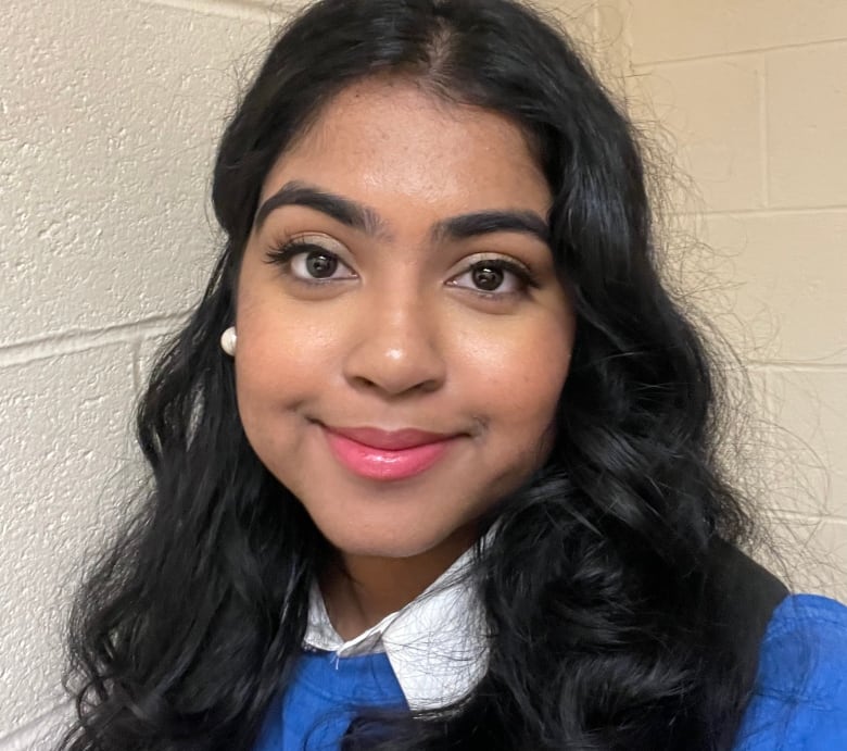 A teenage girl with brown skin and wavy black hair smiles at the camera in a bright blue shirt.