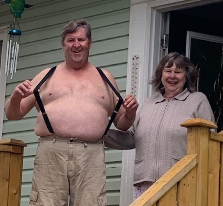 An older man and woman stand next to each other on the front porch of a house. The man doesn't wear a shirt, but suspenders, which he's stretching away from his chest. He has a mischievous smile on his face. The woman next to him is laughing.