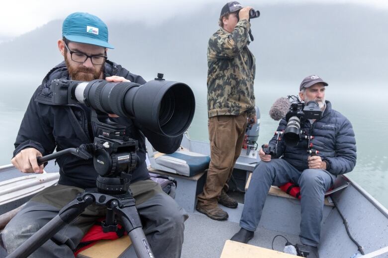 Three men sit in a boat with fog in the background. Two are holding video cameras; one is. holding bnoculars.