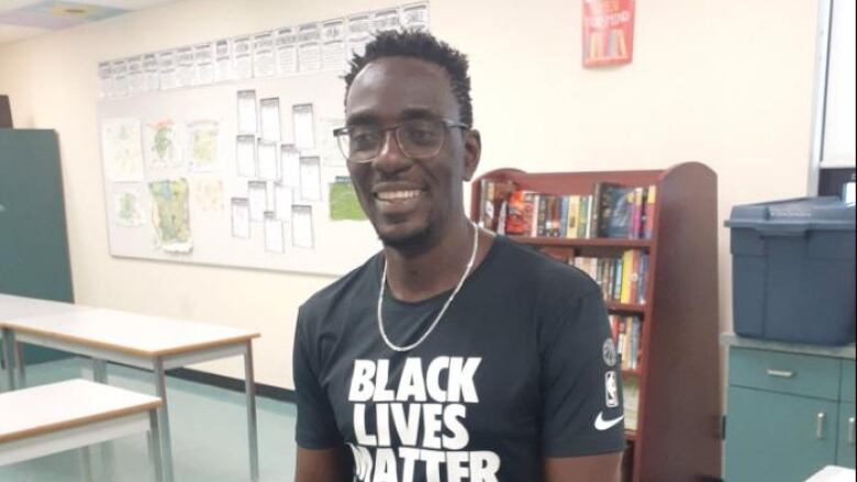 A man in a black T-shirt with words 'Black Lives Matter' sits on a desk in a classroom.