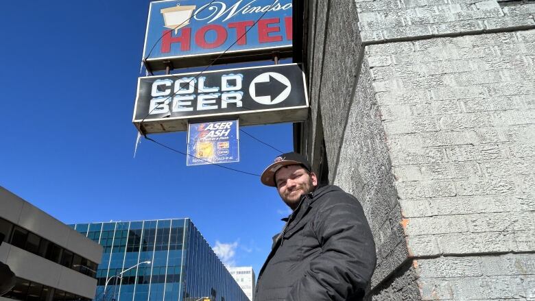 A man wearing a black jacket and baseball hat is standing against a wall. Above him, a sign says Windsor Hotel, and below that, another sign says Cold Beer with an arrow pointing to the right.