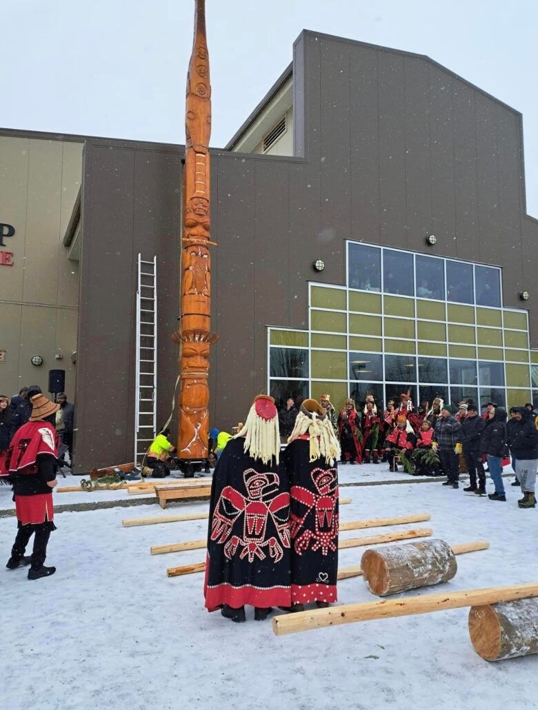 People in Indigenous clothing stand under an erected totem pole that stands in front of a building.