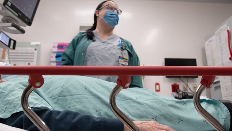 A low camera angle showing a nurse standing over a patient lying on a stretcher, with the monitor for some medical equipment nearby.