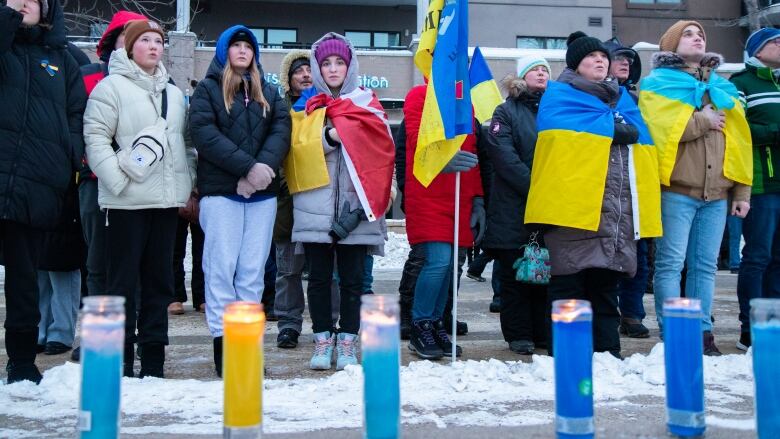 People clad in blue and yellow flags look solemnly at the sky, standing behind a line of blue and yellow candles.