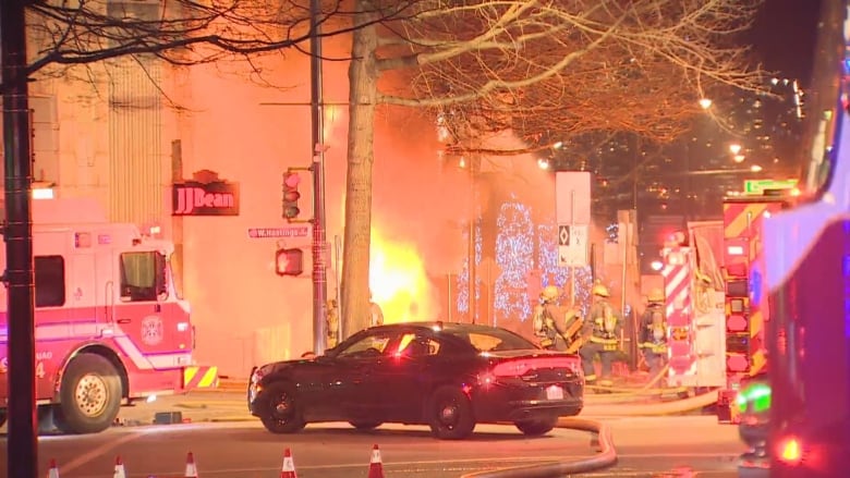 The front end of a fire truck, a black police car and firefighters in full gear are seen working in thick smoke with an orange tint.