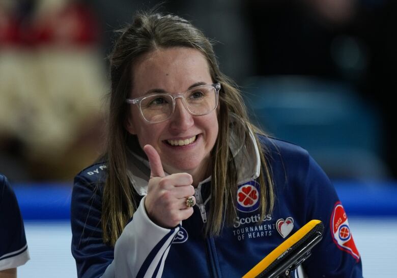 A female curler wearing glasses smiles while giving a thumbs up with her right hand.