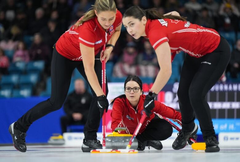 A female curler delivers a rock as two teammates sweep on both sides.