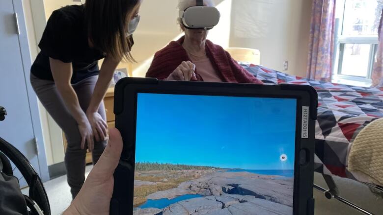 A hand holds a tablet showing a Quebec landscape. In the background, a young woman speaks to an elderly woman who is seated and wearing a virtual reality headset.