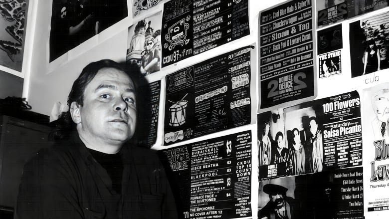 A black-and-white photo shows a man in a small office surrounded by concert posters.