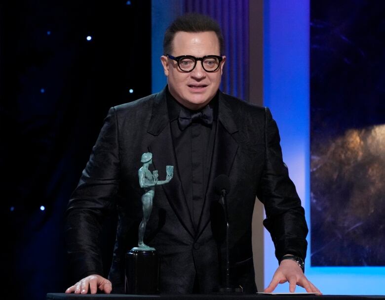 A man in a black tuxedo and glasses stands at a podium to accept an award