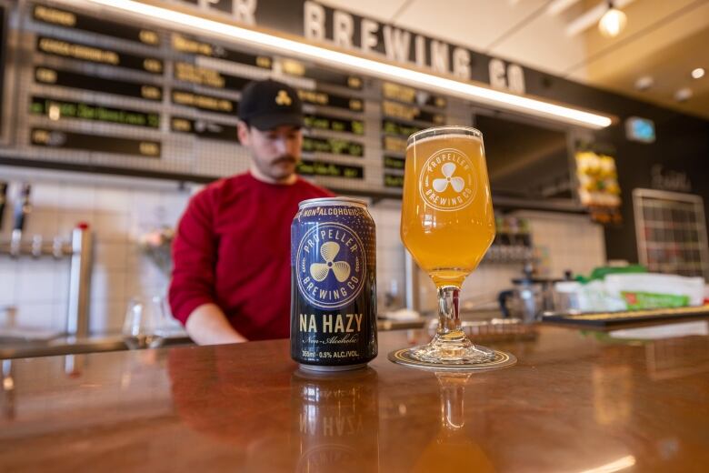 A blue and black beer can with the name NA Hazy is shown on the bar next to a glass that is filled with the beer.