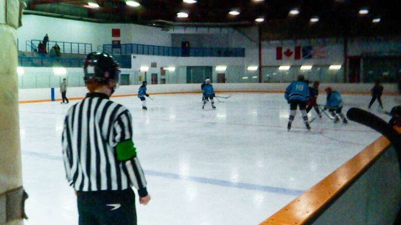 A hockey official wearing a back and white striped shirt watches hockey action at a rink.