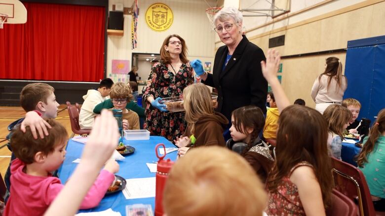 A woman with short grey hair and glasses wears blue plastic gloves and a dark blazer as she talks to a group of children seated at a table in a school gymnasium. Two of the children have their hands raised as if to ask a question.