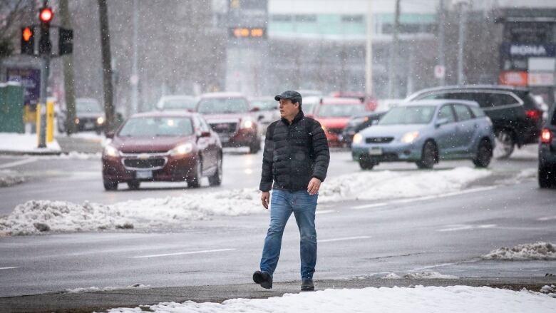 A man walks on the sidewalk as cars drive through snow flurries in the background.