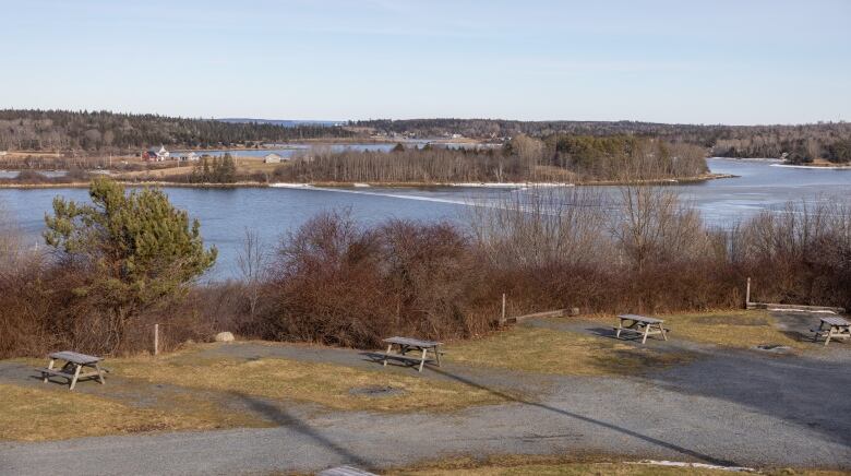 The view shows picnic tables in the foreground and a wooded hill sloping away to a wide expanse of grey-blue water. There's a small peninsula and rolling hills in the background with bare trees beyond and some small homes.