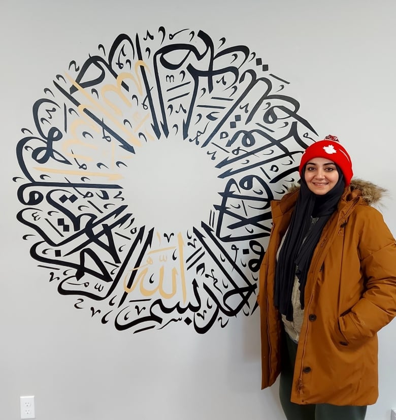 A woman in a toque and coat stands beside a wall mural of of Arabic scripture.