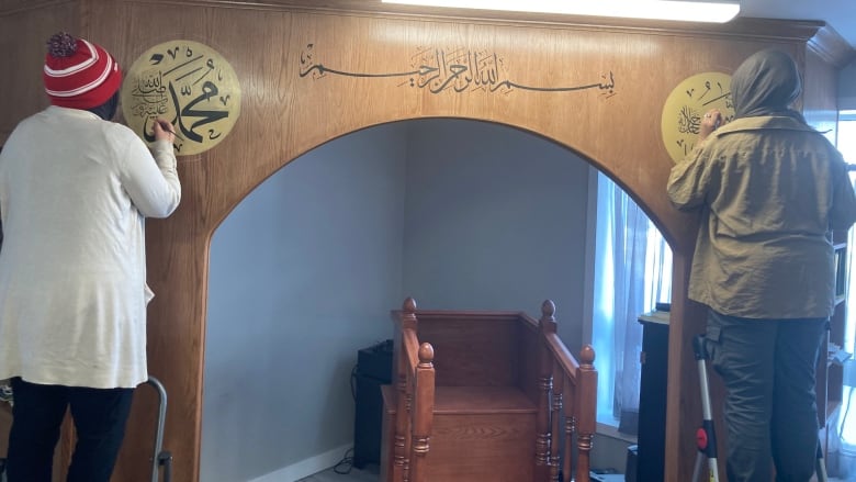 Two people stand on step ladders inside a room, painting Arabic scripture on a wall.