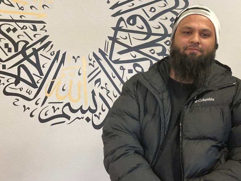 A man in a black winter coat sits in front of a wall decorated with Arabic text.