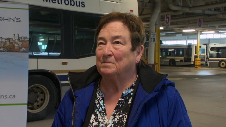 A woman wearing a black shirt and a blue jacket standing inside a large garage. Behind her is a city bus. 