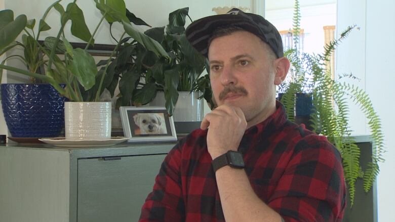 A man wearing a red and black collard shirt and a black baseball cap stands in front of a cabinet with plants on top.