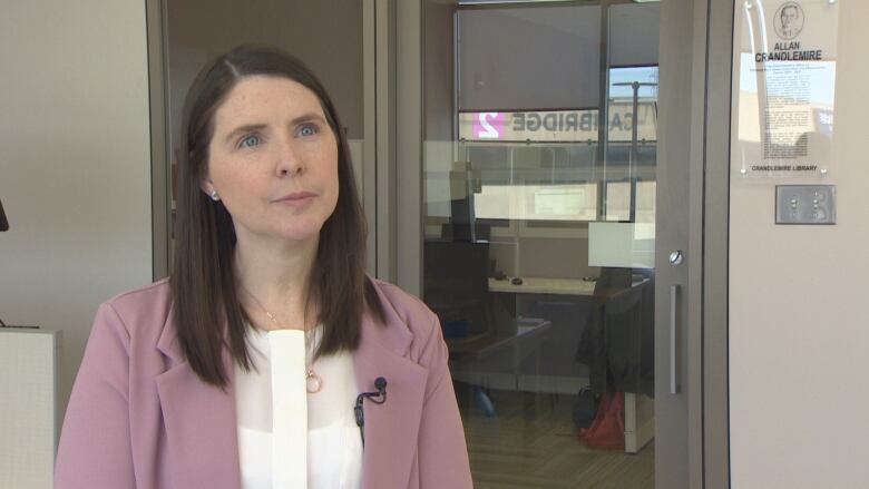 A woman with blue eyes and medium-length brown hair wears a white shirt with a purple blazer. She is standing in front of a glass door.