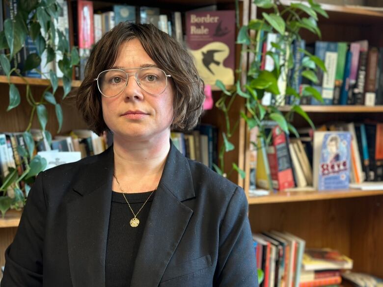 A woman with jaw-length hair and glasses poses in a dark shirt and dark blazer in front of a book shelf.