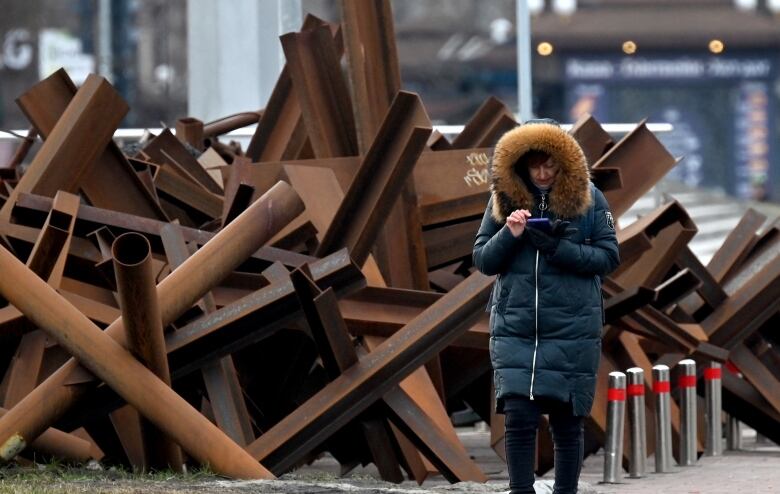 A woman wearing a winter jacket looks down at her cell phone as she walks past piles of large steel beams and pipes on a city street.