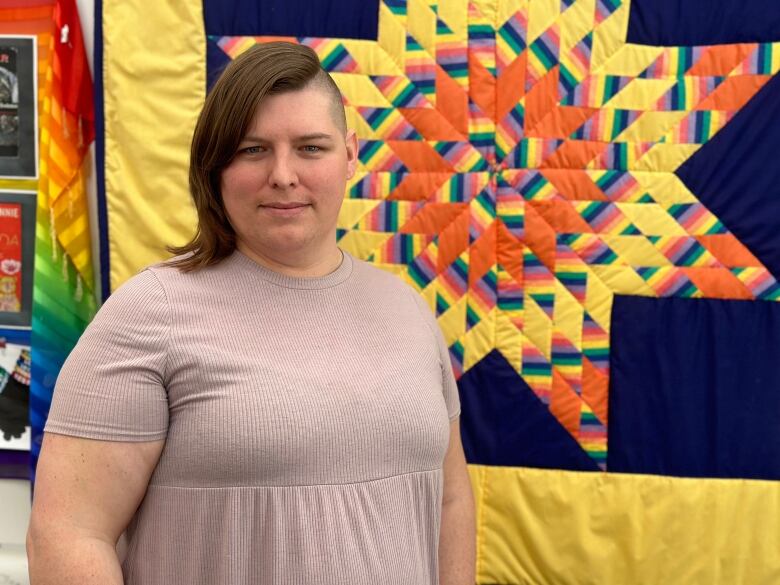 A person in a light-coloured t-shirt poses in front of a rainbow mosaic quilt hanging on a wall.