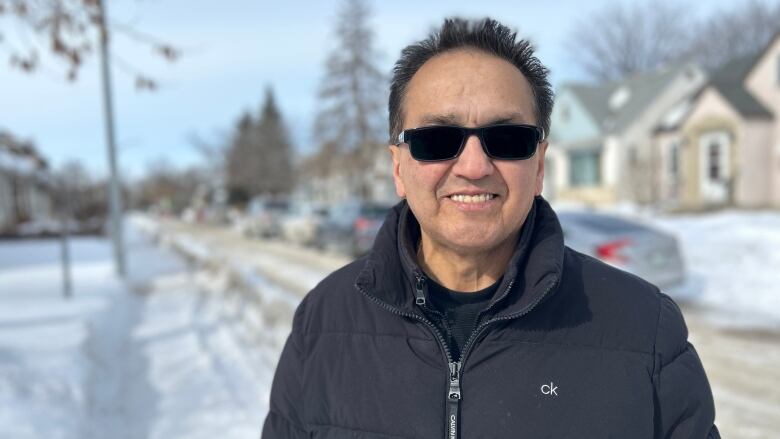 A man wearing a black jacket and sunglasses is standing on a sidewalk, with a snowy street behind him.