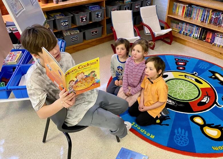 An older student on a chair reads a book to three younger students.