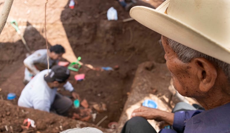 A seated man looks on as archaeologist recover bones from the red soil.