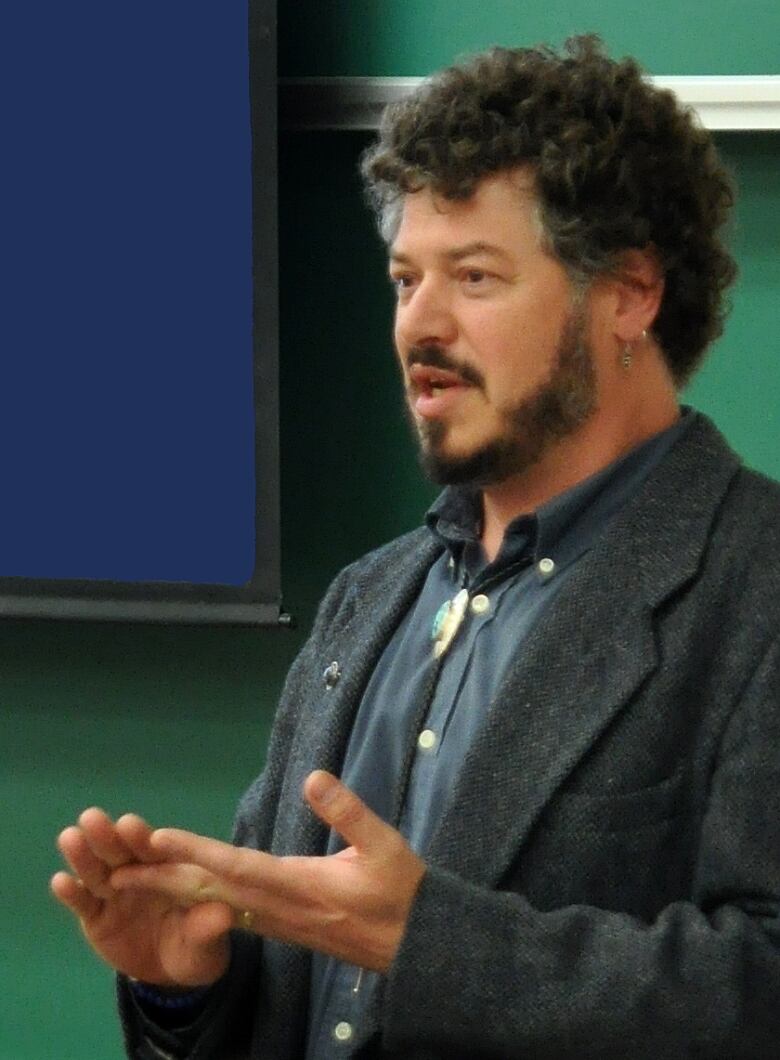 A bearded man in a button-down shirt and blazer speaks at an indoor public event.