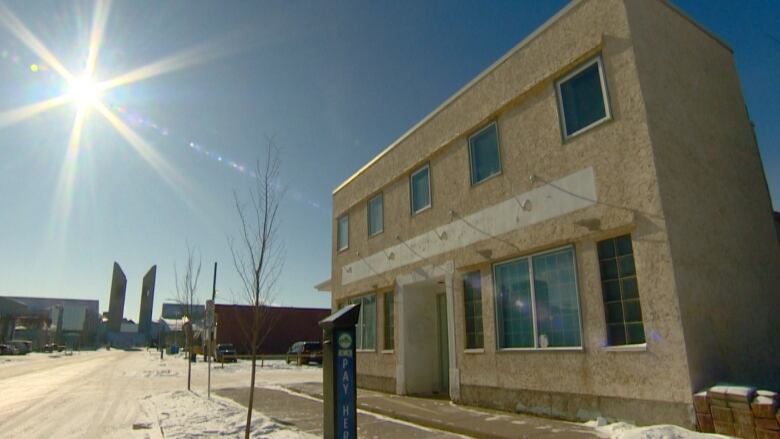 A two storey white building with a dozen windows and door set against a snowy street with MacEwan University in the background as the sun shines.