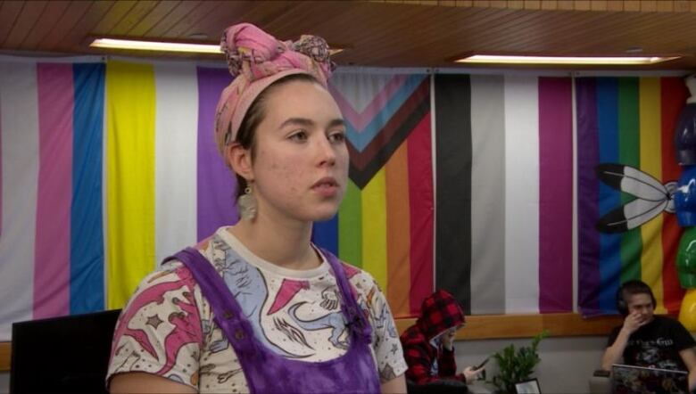 A woman with a pink headband stands in front of a rainbow flag wall.