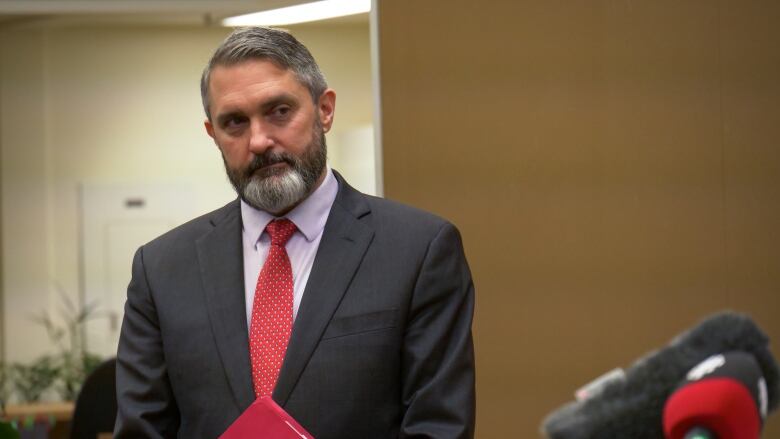 A bearded man in a suit stands holding a red folder, with microphones visible in the foreground.