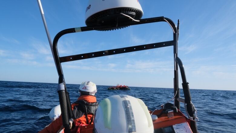 The helmeted heads of two people on a boat are shown in the foreground, as another boat, loaded with people wearing orange life-jackets, is seen in the distance.