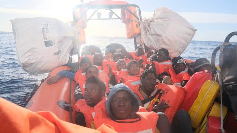 A large group of people wearing life-jackets are crowded in an orange inflatable raft at sea. Large white bags of supplies hang from the boat.