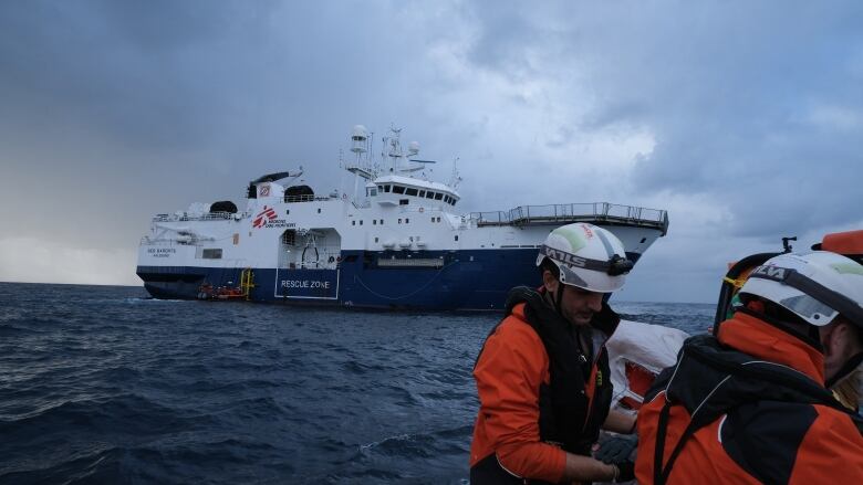 A large blue and white boat is seen at sea, while in the foreground, two men dressed in white helmets and orange jackets work in a smaller boat.