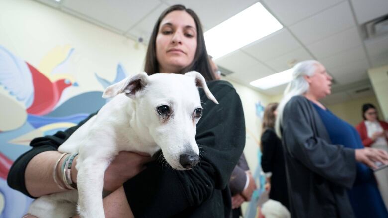 A woman holds a small white dog in her arms as another woman speaks into a microphone behind her. 