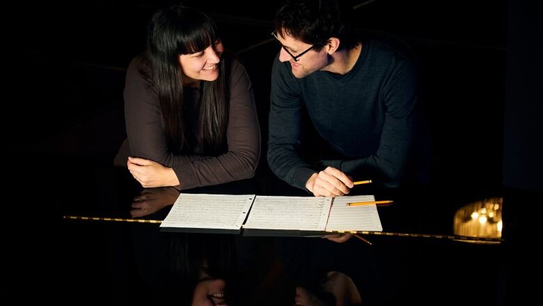 Two people sit side by side leaning over a grand piano with sheet music scattered in front of them, smiling at each other.