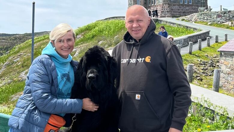 A man and a woman look at the camera and a black Newfoundland dog are in between them. 