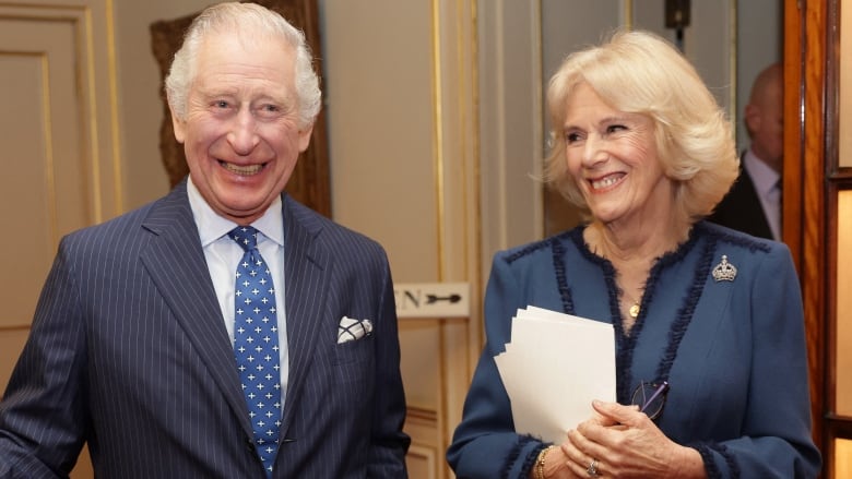 King Charles and Camilla standing in a room together dressed in blue. Camilla is holding papers.