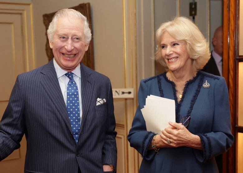 King Charles and Camilla standing in a room together dressed in blue. Camilla is holding papers.